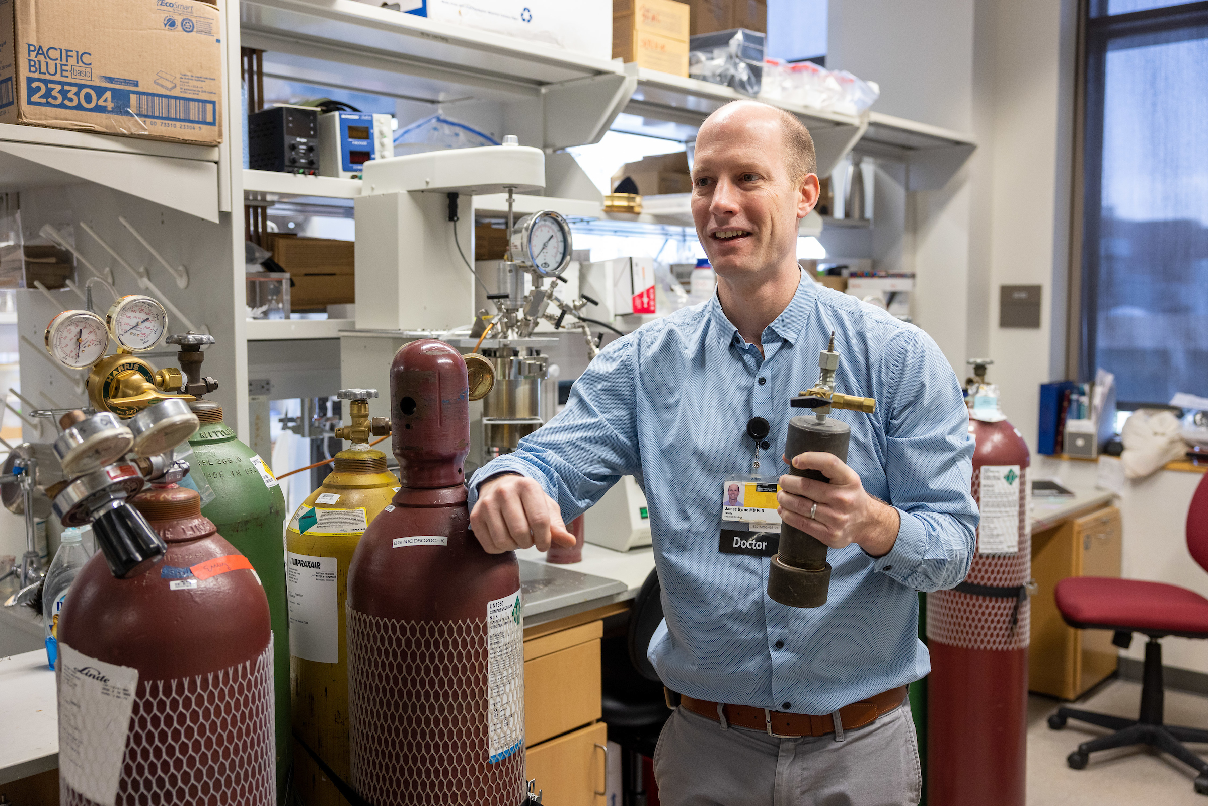 Dr. James Byrne holds the modified whipping siphon in his lab at the University of Iowa. Around him are large tanks of pressurized gas.