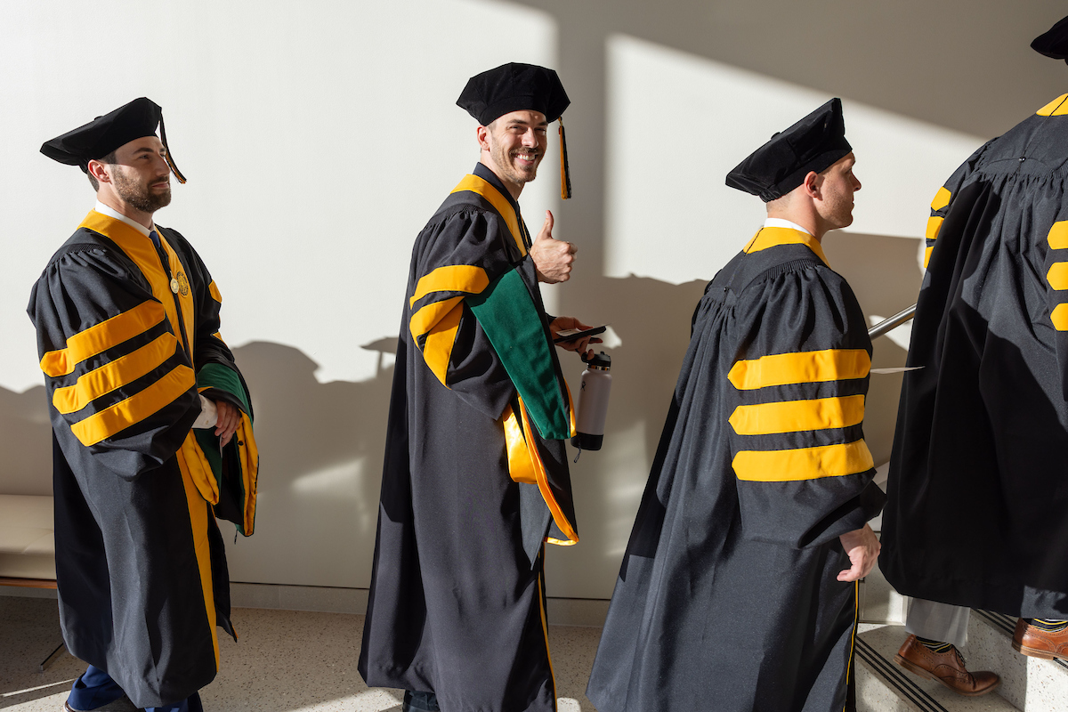 Four students in their ceremonial robes walk down a sunny hallway in Hancher Auditorium. One gives a thumbs-up to the camera.