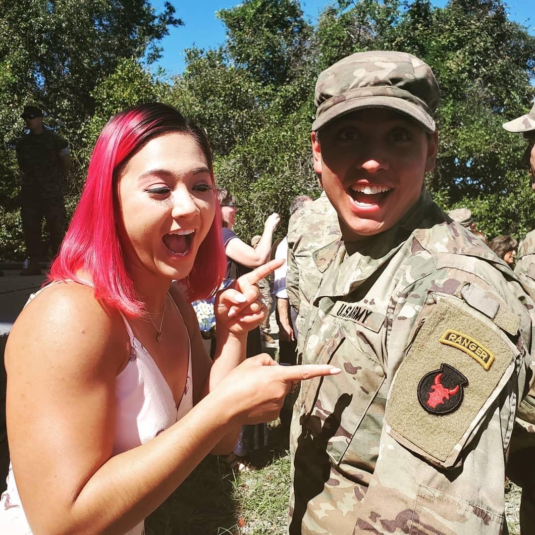 Daniel, in his uniform, stands with his wife Amy. Both are smiling and Amy is pointing at the insignia on his shoulder that designates Daniel as an Army Ranger.