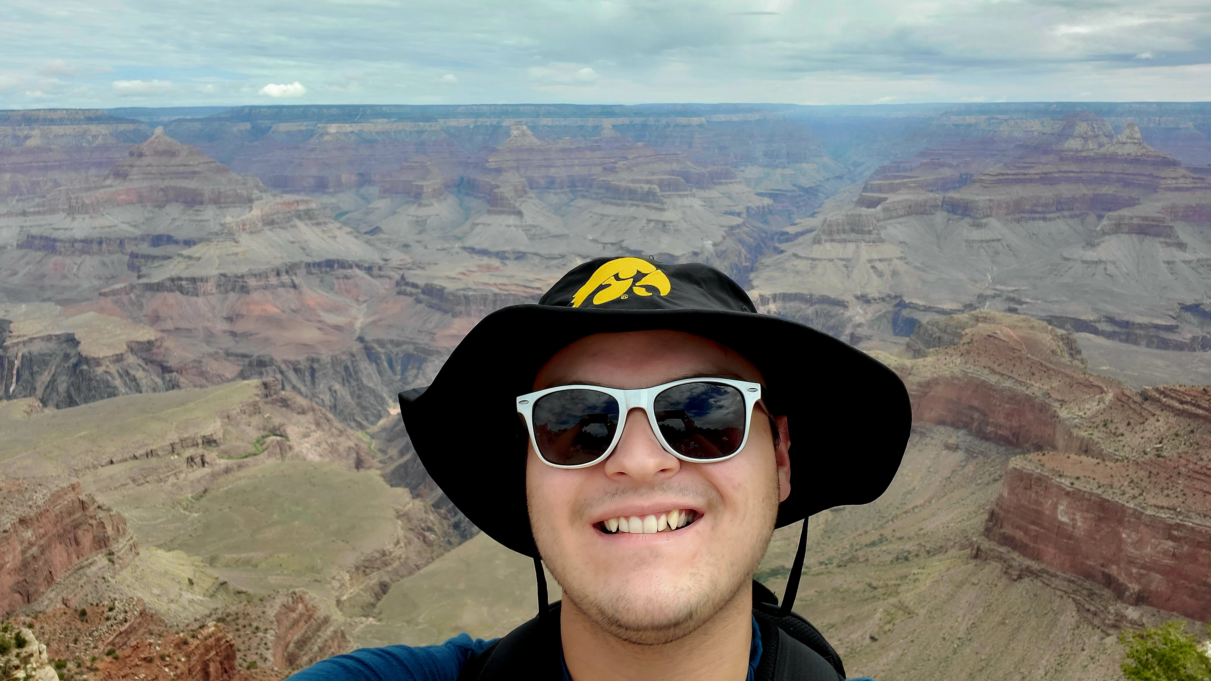 Selfie of Travis Fischer with a mountainous landscape in the background. He is wearing sunglasses and an Iowa Hawkeyes hat.