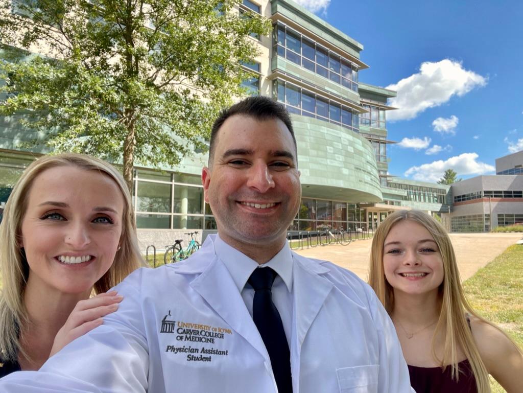 Selfie of John Hanshew with his wife and daughter. He is wearing his PA white coat and the medical campus is visible in the background..