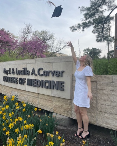 Ellie Peters throwing her graduation cap in the air next to the Carver College of Medicine sign.