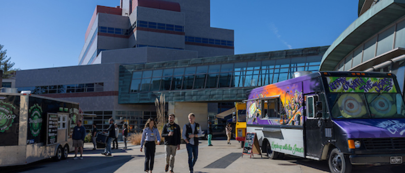 Three students walk in a campus pedestrian area that contains two food trucks.